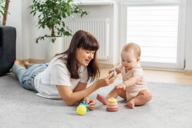 Gülümseyen anne halının üzerinde yatıyor. Bebeğine rehberlik ediyor. Yüzük istiflemeyi, erken öğrenmeyi, eğlenmeyi ve aile bağlarını teşvik ediyor..