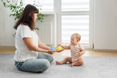 Meraklı bebek halıya oturur ve annesinin ellerinde renkli duyusal toplara uzanır, neşeli aile bağlarını ve erken öğrenmeyi gösterir.