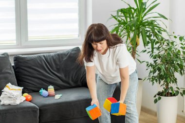 Parlak bir oturma odasında renkli çocuk oyuncaklarını almak için eğilen bir kadın günlük ebeveynlik rutinini gösteriyor.