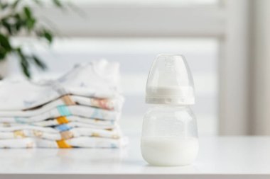 Süt dolu bir bebek şişesi beyaz bir masanın üzerine koyuldu. Renkli, katlanmış, yumuşak ışıkta bebek kıyafetlerinin yanına..
