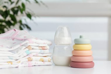 Bebek sütü şişesi, pastel yüzüklerin arasına yerleştirilmiş ve bembeyaz bir masanın üzerinde düzgünce katlanmış bebek kıyafetleri, yumuşak ışıklı bir odada..