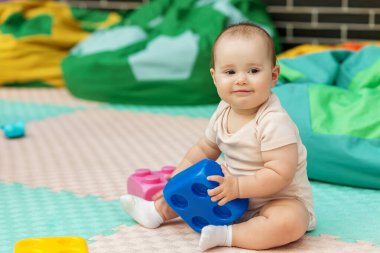Kapalı alanda oyun hasırının üzerinde oturan mutlu bebek renkli bir yapı taşı tutuyor. Ebeveynlik, kreş ya da çocukluk kavramları için mükemmel bir görüntü..