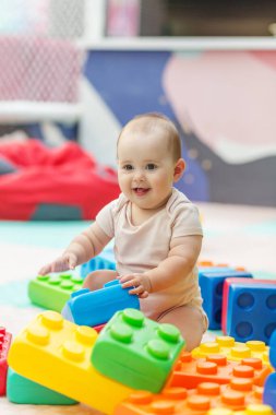 Anaokulunun oyun sahasında, parlak plastik bloklarla çevrili yumuşak bir mindere oturan mutlu bebek. Ebeveynlik ve eğitim kullanımı için ideal.