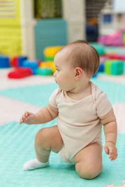 Bej tulumlu sevimli bebek, renkli oyuncaklarla çevrili yumuşak bir oyun paspasına oturmuş, meraklı bir şekilde oyun odasına bakıyor..