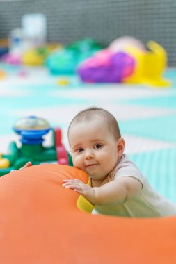 Şirin bebek, kapalı alandaki çocukların oyun ortamında top oyuncağına uzanırken turuncu yumuşak oyun yapısına yaslanıyor.
