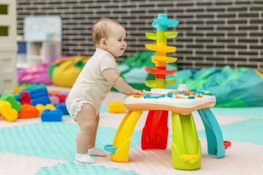 Anaokulunda renkli bir oyun masasında oynayan gülümseyen bebek oyuncakları keşfediyor ve ilk motor becerilerini geliştiriyor..