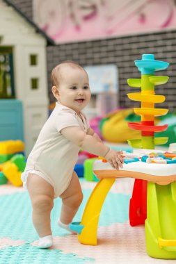 Gülümseyen bebek kapalı bir oyun odasında renkli oyuncaklarla çevrili eğitici bir aktivite masasıyla ayakta duruyor ve oynuyor..
