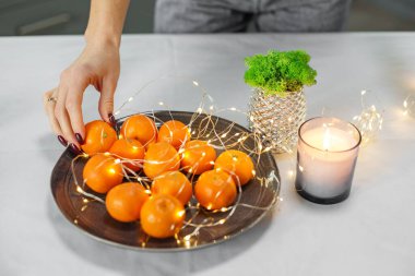 Peri ışıklarıyla süslenmiş bir tabakta taze portakallara uzanan bir elin yakın çekimi, yanan bir mumun yanında..