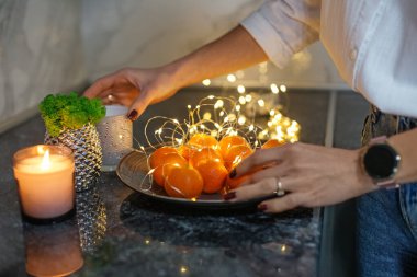 Yakın plan çekimde taze portakallı dekoratif bir tabak ve yanan bir mumun yanındaki tezgahın üzerinde parlayan peri ışıkları düzenleniyor..