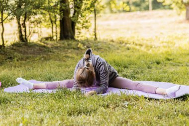 Genç bir kız çimenli bir parkta yoga minderinin üzerinde geniş bir ayrılık içinde, esneklik ve denge pratiği yapıyor..