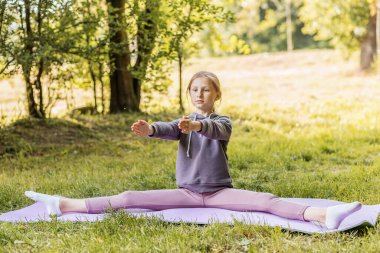 Spor kıyafetli genç bir kız ağaçlarla çevrili güneşli bir açık hava parkında yoga minderinde tam bir bölünme sergiliyor..