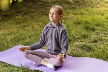 Gülümseyen ergen bir kız yeşil bir parkta mor bir yoga minderinin üzerinde oturmuş meditasyon ve açık havada farkındalık yapıyor..