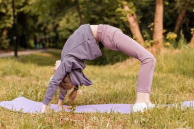 Genç bir kız, parktaki yoga minderinde, esneklik, güç ve açık hava ortamında odaklanma göstererek geri dönüş yapıyor..