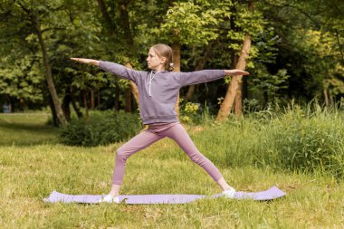 Odaklanmış kız, doğadaki yoga minderinin üzerinde duruyor. Açık hava antrenmanı sırasında gücü, dengeyi ve farkındalığı teşvik ediyor..