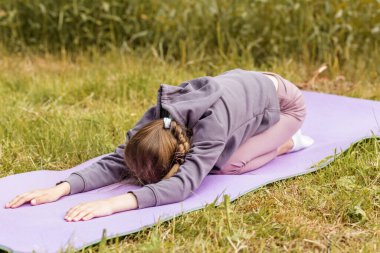 Genç bir kız yoga minderinde çocuk pozu pratiği yapıyor. Rahatlama, farkındalık ve açık hava ortamında iç huzuru gösteriyor..