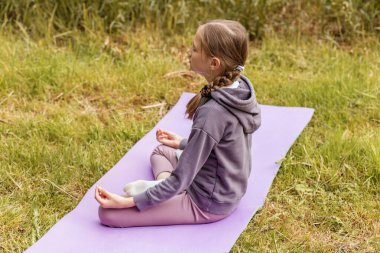 Genç bir kız parkta yoga minderinde bacak bacak üstüne atmış, rahatlama ve sakinlik için meditasyon ve farkındalık yapıyor..