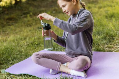 Genç bir kız parkta yoga minderinde bacak bacak üstüne atarak oturur. Egzersiz sonrası su şişesini açarken gülümser..