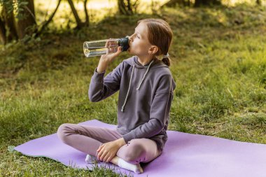 Genç bir kız parkta yoga minderinde bacak bacak üstüne atarak oturur, açık havada egzersiz yaptıktan sonra şişeden su içer..
