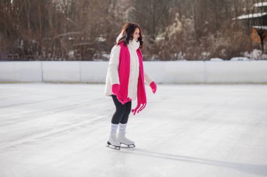 Kış kıyafetleri içinde, açık havada güneşli bir kış günü buz pateni pistinde açık pembe bir atkıyla buz pateni yapan kendine güvenen bir kadın..