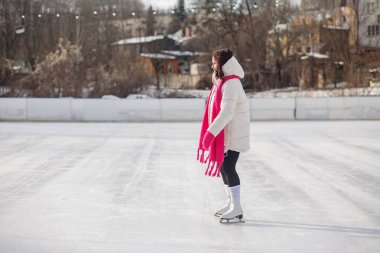 Sıcak kış kıyafetleri ve pembe aksesuarlı bir kadın, soğuk ve güneşli bir günde buz pistinde paten kayıyor..
