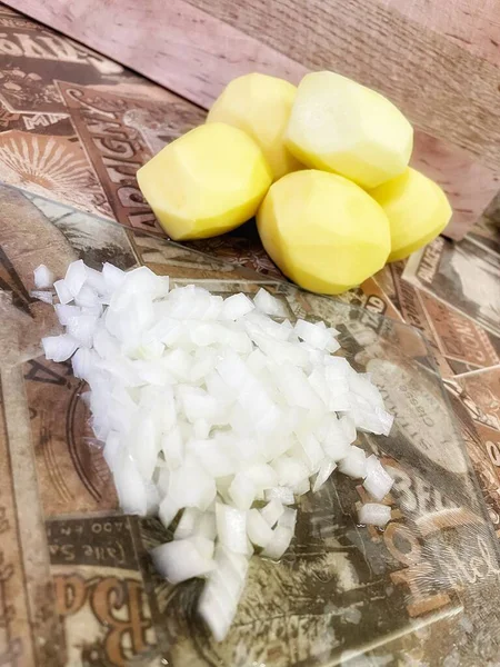Mutfak masasında çiğ, soyulmuş büyük patatesler ve ince doğranmış soğanlar var; 