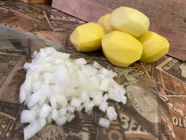 Mutfak masasında çiğ, soyulmuş büyük patatesler ve ince doğranmış soğanlar var..