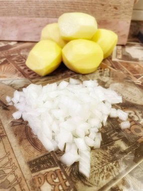 Mutfak masasında çiğ, soyulmuş büyük patatesler ve ince doğranmış soğanlar var..
