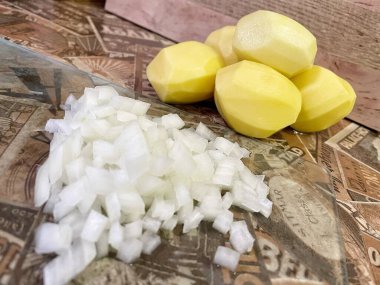 Mutfak masasında çiğ, soyulmuş büyük patatesler ve ince doğranmış soğanlar var..