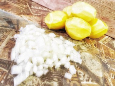 Mutfak masasında çiğ, soyulmuş büyük patatesler ve ince doğranmış soğanlar var..