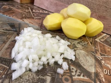 Mutfak masasında çiğ, soyulmuş büyük patatesler ve ince doğranmış soğanlar var..