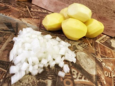 Mutfak masasında çiğ, soyulmuş büyük patatesler ve ince doğranmış soğanlar var..