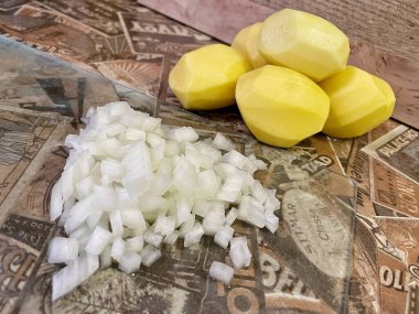 Mutfak masasında çiğ, soyulmuş büyük patatesler ve ince doğranmış soğanlar var..