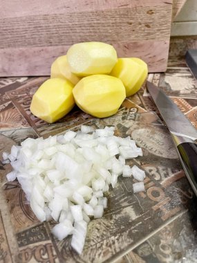 Mutfak masasında çiğ, soyulmuş büyük patatesler ve ince doğranmış soğanlar var..