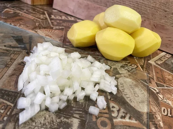 Mutfak masasında çiğ, soyulmuş büyük patatesler ve ince doğranmış soğanlar var..