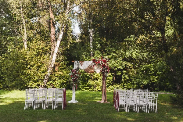 Wedding ceremony in rustic style decorated with red flowers and textile with white chairs in forest