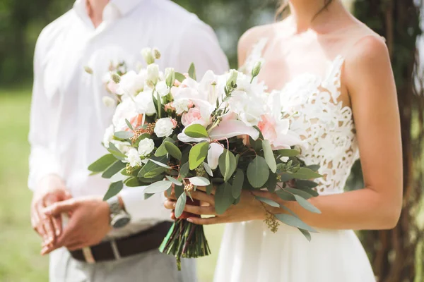 Gentle wedding bouquet with lily, pink roses and greens in loving couple hands