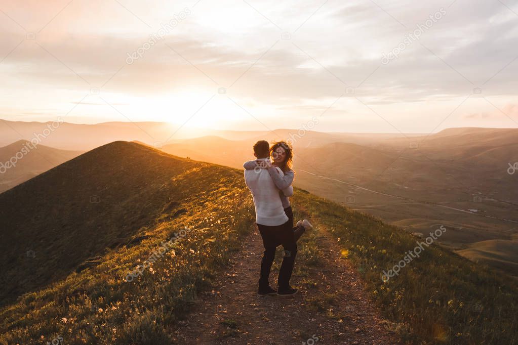 Happy couple hugging and kissing at sunset with amazing mountains view. Warm evening sun light