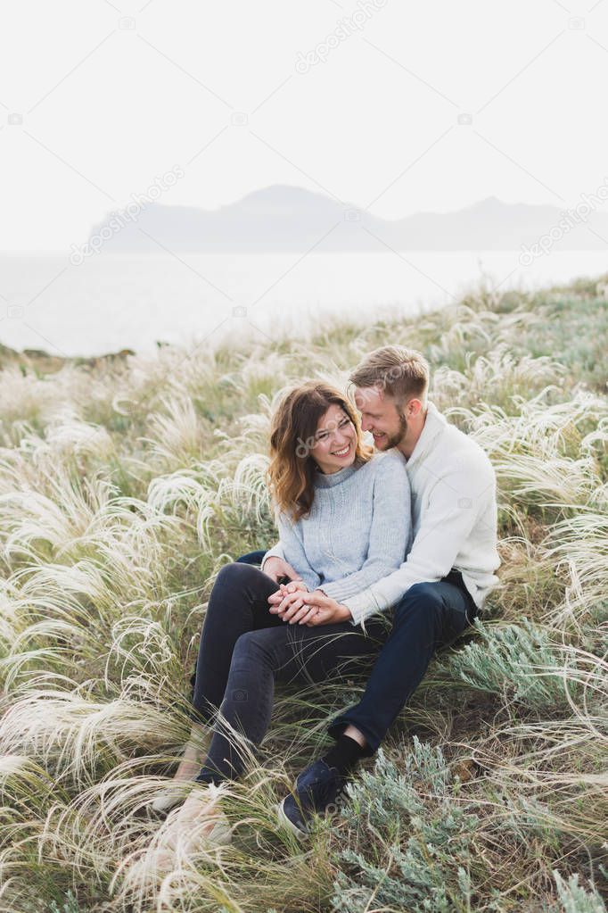 Happy young loving couple sitting in feather grass meadow, laughing and hugging, casual style sweater and jeans