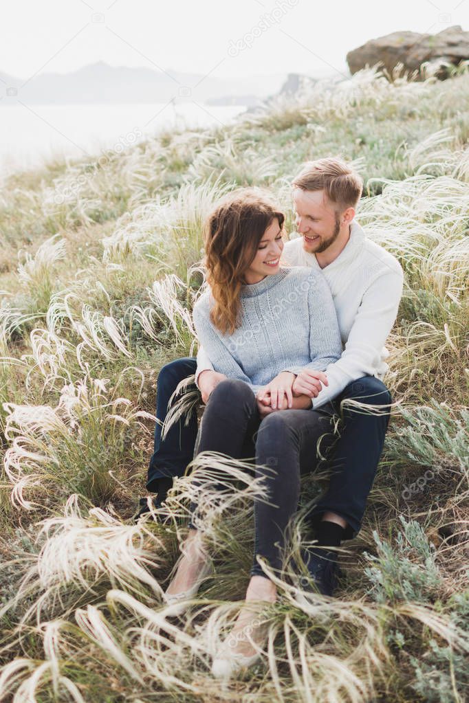 Happy young loving couple sitting in feather grass meadow, laughing and hugging, casual style sweater and jeans