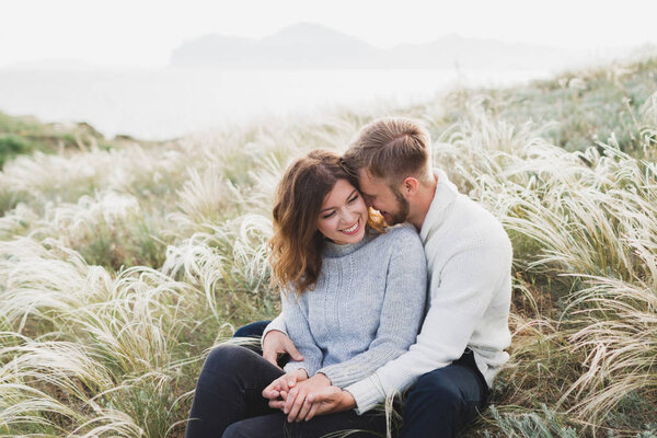 Счастливая молодая любящая пара, сидящая на лугу из перьев, смеющаяся и обнимающаяся, свитер и джинсы повседневного стиля
