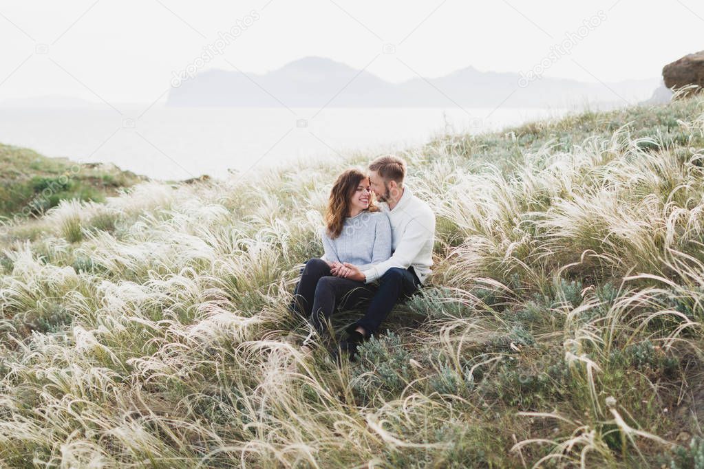 Happy young loving couple sitting in feather grass meadow, laughing and hugging, casual style sweater and jeans