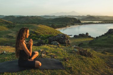 Kadın güneşin doğuşunda dağ ve okyanus manzarası ile tek başına yoga yapıyor. 