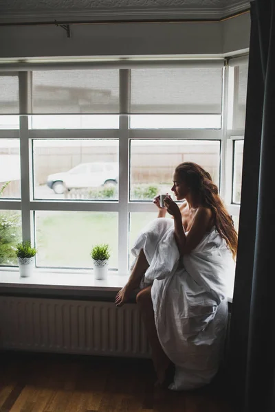 Yung žena pokrytá dekou sedí na parapetu a těší — Stock fotografie