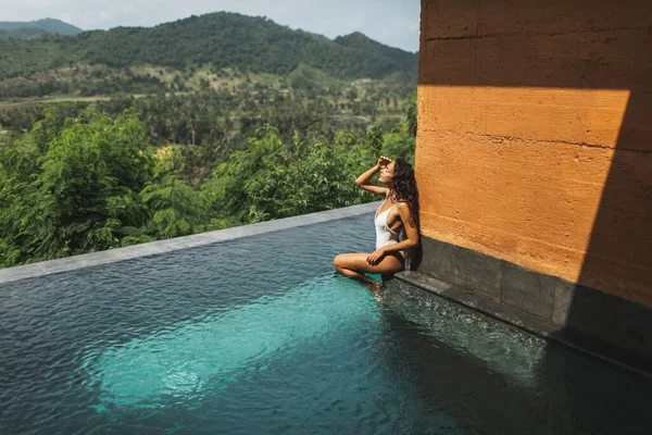 Beautiful young woman sunbathing and relaxing on edge private pool with amazing jungle and mountain view. Summer vacation, inspiration travel. White swimsuit.