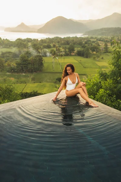 Dream journey in Asia. Woman sitting on edge luxury infinity pool an enjoying amazing mountain, rice terraces and coastline view at sunset. Inspiration travel destination.
