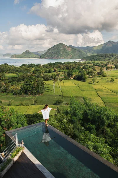 Woman in white tunic walking by the edge infinity pool with awesome view of rice terraces, mountains and ocean coast. Aerial view. Vacations in Asia.