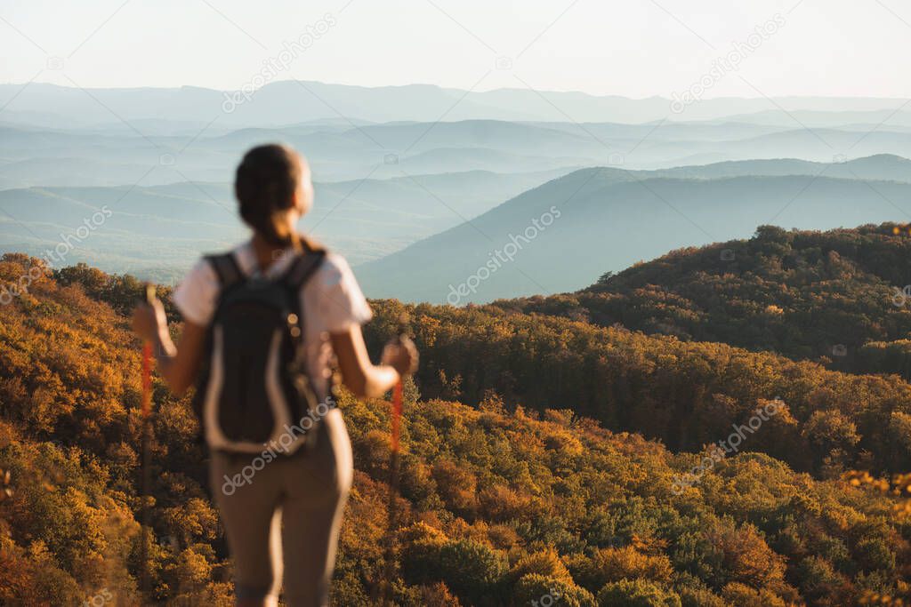 Rear view of woman enjoying autumn layered mountain landscape. Orange autumn forest and blue hills in mist on horizon. Beauty in nature, empty  place on background.