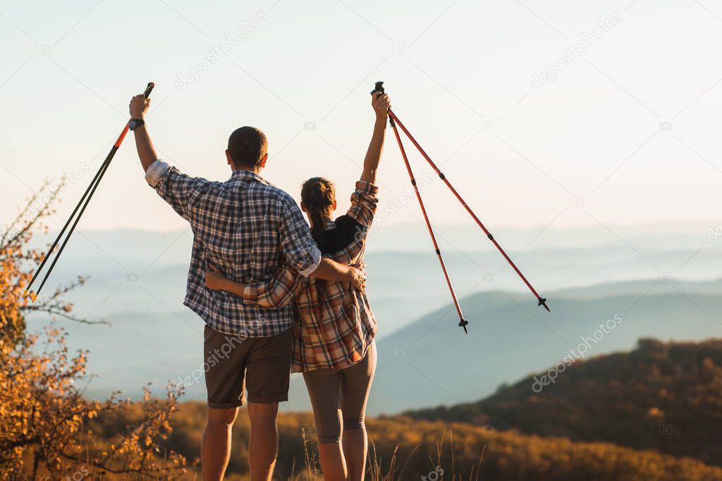 Couple happy to climb top of mountain and hold trekking sticks. Nordic walking outdoors, view from behind. Travel lifestyle. Success goal achievement.