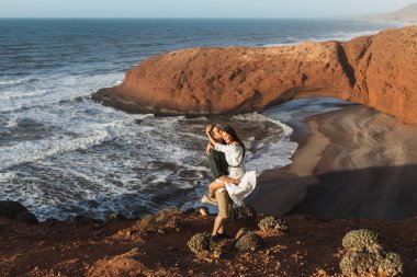Young happy smiling couple hugging in love on background of famous arch on Legzira beach in Morocco. Sidi Ifni landscape. Popular moroccan landmark. clipart
