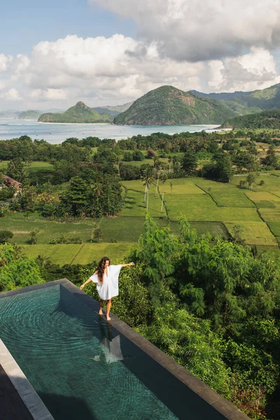 Woman in white tunic walking by the edge infinity pool with awesome view of rice terraces, mountains and ocean coast. Aerial view. Vacations in Asia.